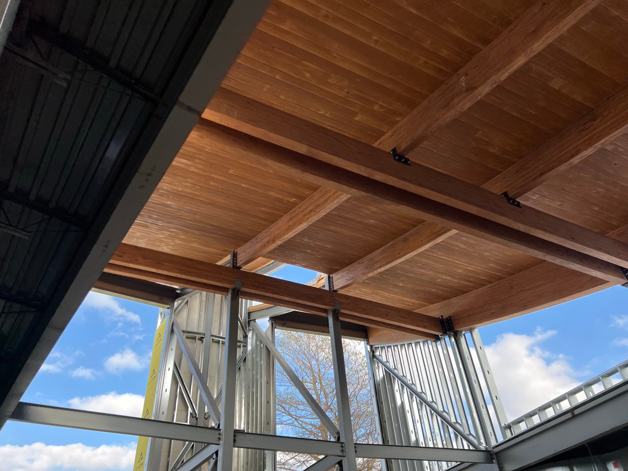 Construction site framing with exposed wooden beams and metal studs against a blue sky, showing an unfinished building interior/roof space.
